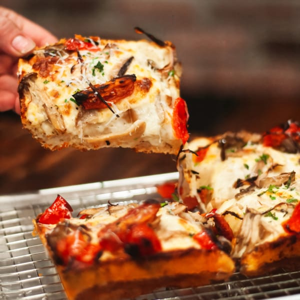 Hand lifting a square slice of Croccante Artisan Pizza with mushrooms, tomatoes, and cheese.