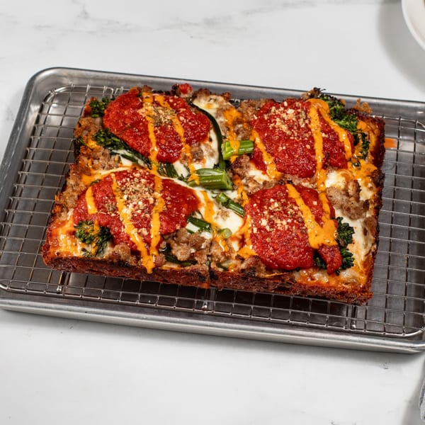 A rectangular Croccante Artisan Pizza with red sauce, cheese, sausage, and green vegetables on a metal rack.