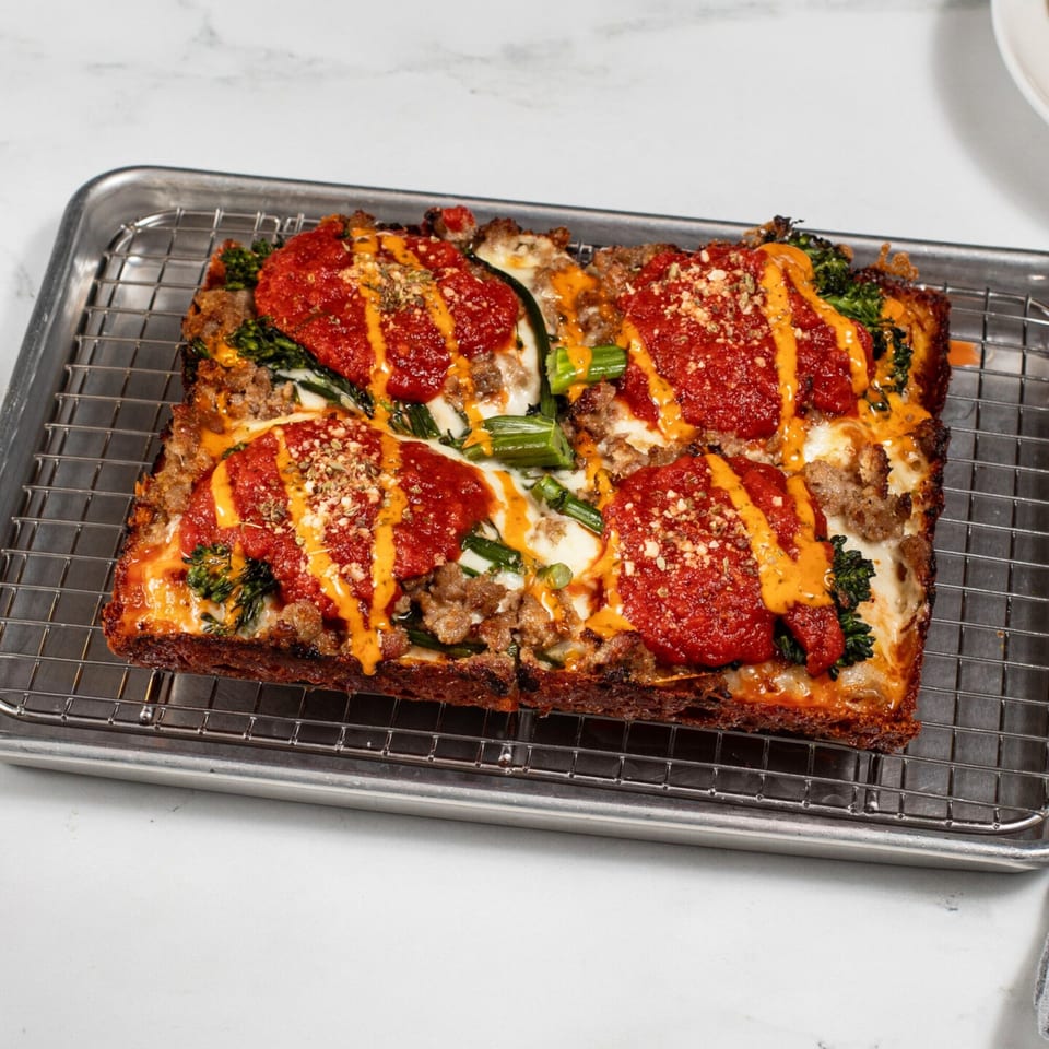 A rectangular Croccante Artisan Pizza with red sauce, cheese, sausage, and green vegetables on a metal rack.