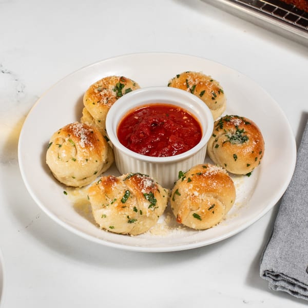 Garlic knots from CROCCANTE ARTISAN PIZZA, sprinkled with parmesan and parsley, surrounding a marinara dipping sauce.