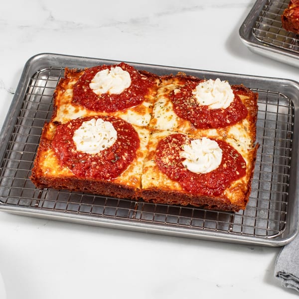 A rectangular Detroit-style pizza with four dollops of ricotta cheese and red sauce on a cooling rack.