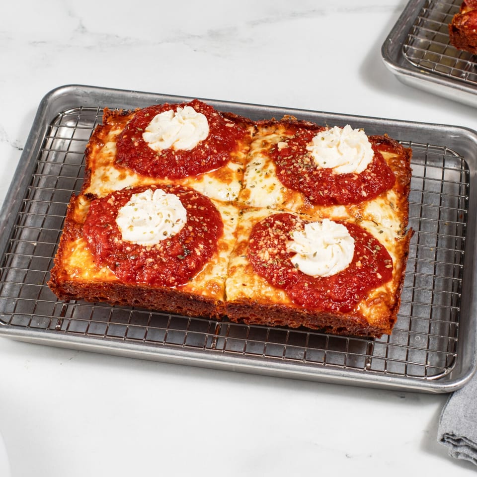 A rectangular Detroit-style pizza with four dollops of ricotta cheese and red sauce on a cooling rack.