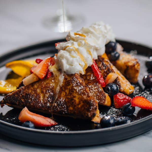 Golden French toast triangles topped with whipped cream, berries, and a drizzle of syrup on a dark plate.