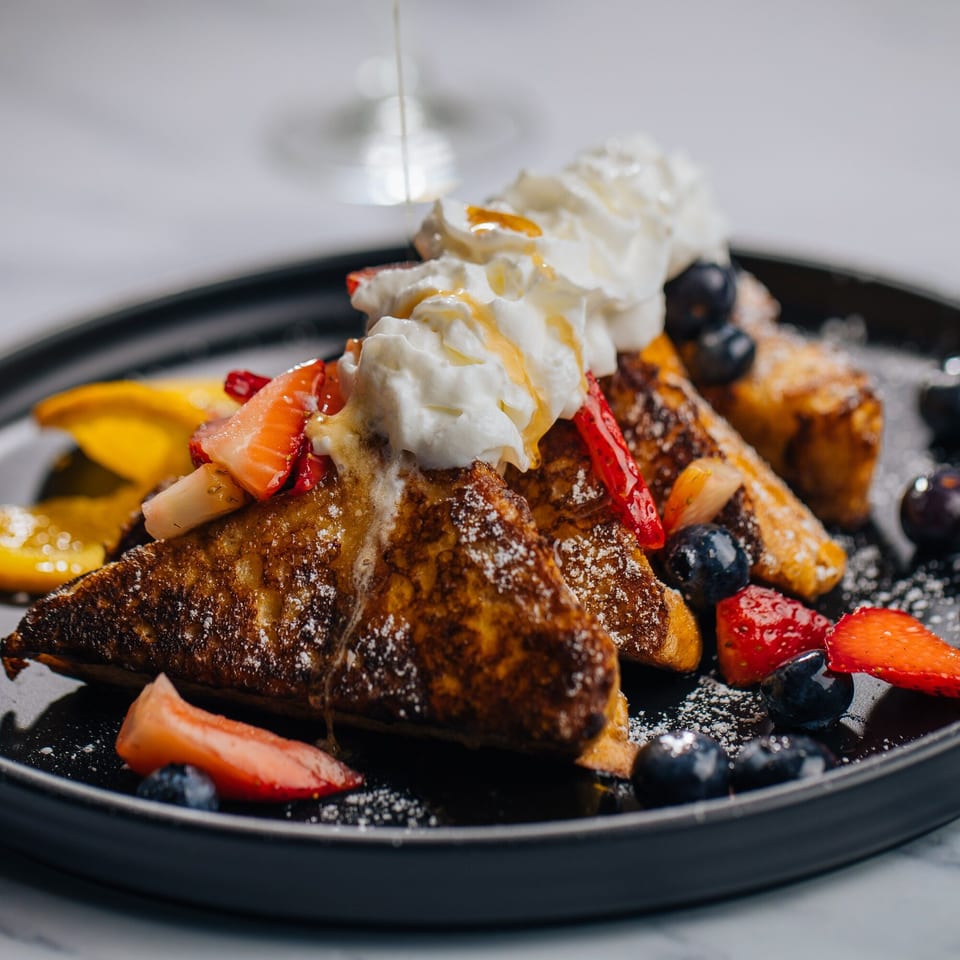 Golden French toast triangles topped with whipped cream, berries, and a drizzle of syrup on a dark plate.