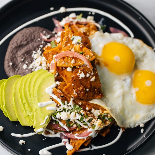 Chilaquiles with two fried eggs, black beans, avocado slices, and crema on a black plate.