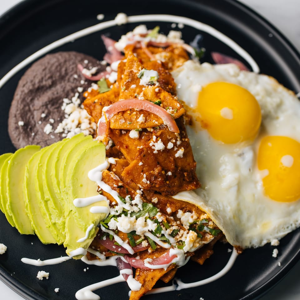 Chilaquiles with two fried eggs, black beans, avocado slices, and crema on a black plate.