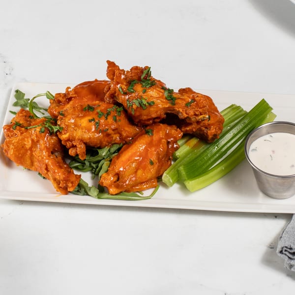 Buffalo chicken wings with celery sticks, arugula, and a side of ranch dressing on a white plate.