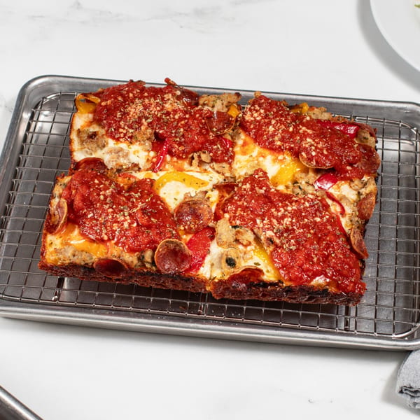 Detroit-style pizza with pepperoni, sausage, bell peppers, and tomato sauce on a cooling rack.