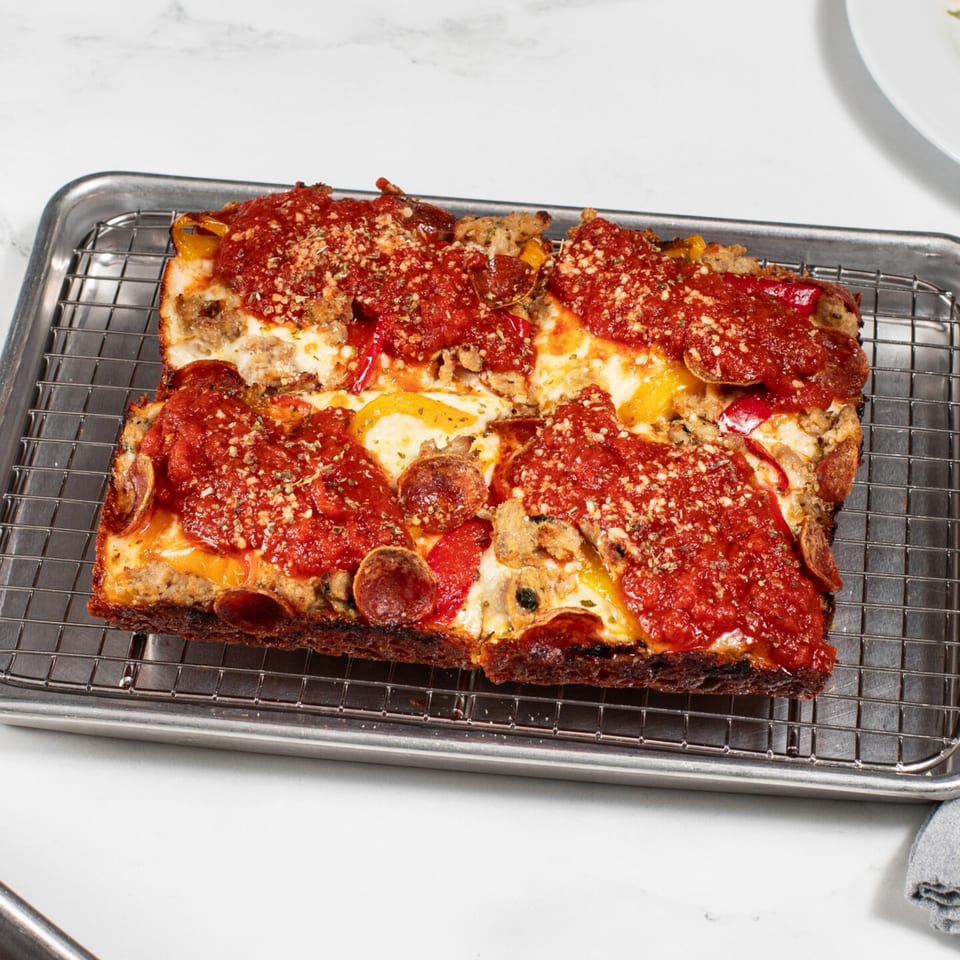 Detroit-style pizza with pepperoni, sausage, bell peppers, and tomato sauce on a cooling rack.