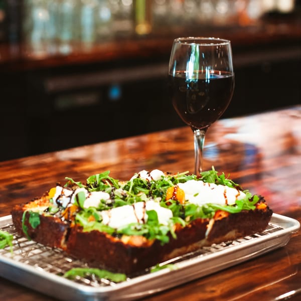 CROCCANTE ARTISAN PIZZA's square pizza with arugula, burrata, and balsamic glaze, next to a glass of red wine.