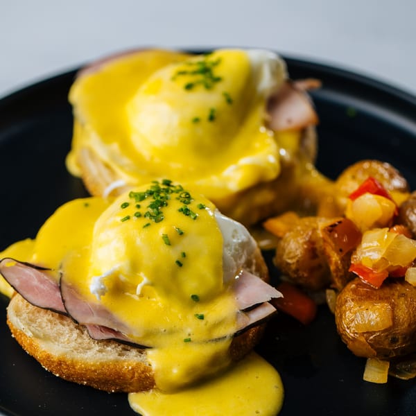 Two eggs benedict with ham and hollandaise sauce, garnished with chives, served with roasted potatoes.
