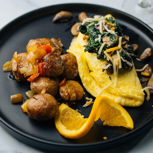 A breakfast plate with an omelet topped with spinach and cheese, roasted potatoes, and an orange slice.