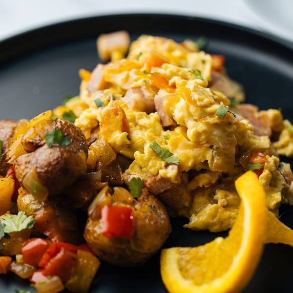 Scrambled eggs with diced ham, roasted potatoes, red peppers, and a fresh orange slice.